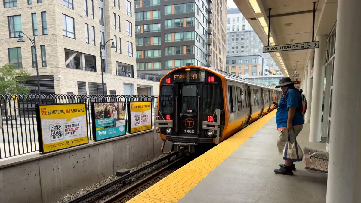 A person looking at his smartphone on a station platform as an orange and silver train approaches, with tall buildings in the background.